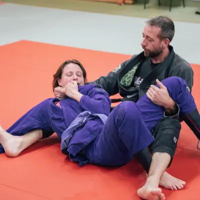 Zwei Personen trainieren Jiu-Jitsu auf einer roten Matte, ein Mitglied des Vereins TGL demonstriert eine Technik.