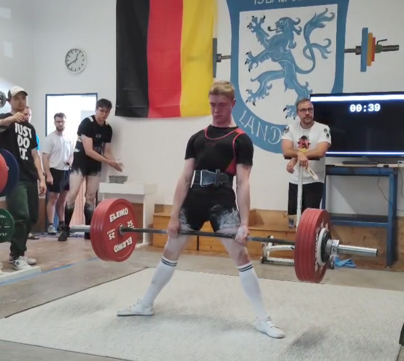 Ein Gewichtheber hebt eine Langhantel bei einem Wettkampf der Turngemeinde Landshut, während Zuschauer applaudieren.