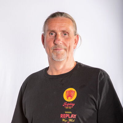 Ein Mann mit grauem Bart trägt ein T-Shirt mit bunten Logos und repräsentiert den Sportverein Turngemeinde Landshut.