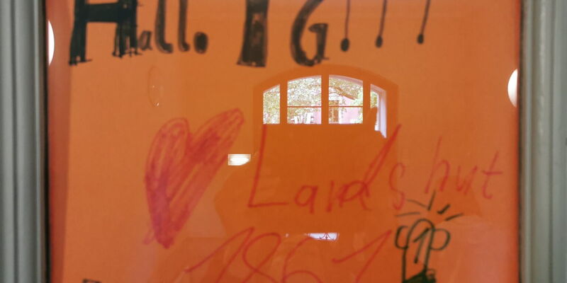 Ein orangefarbenes Plakat begrüßt die Turngemeinde Landshut mit fröhlichen Zeichnungen und Schriftzügen an einer Glastür.