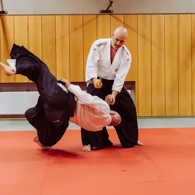 Zwei Männer üben eine Aikido-Technik in einer Turnhalle des Sportvereins Landshut auf einem roten Mattenboden.