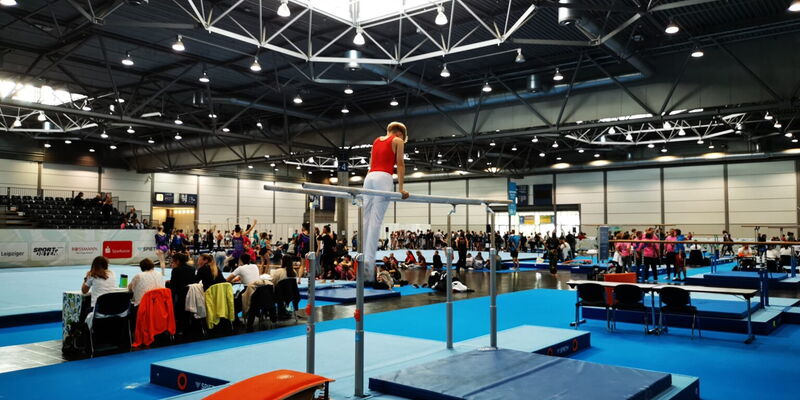 Ein Turner der Turngemeinde Landshut zeigt eine Übung am Barren in einer belebten Sporthalle.