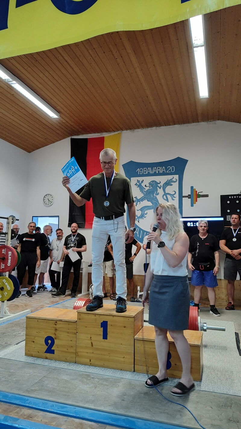 Ein Mann steht mit Medaille auf dem Siegerpodest bei einem Turngemeinde-Wettbewerb in Landshut, während eine Frau spricht.