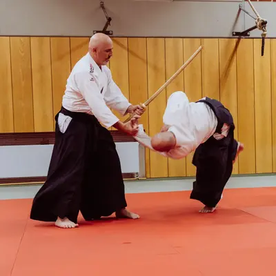 Zwei Männer üben Aikido-Techniken mit einem Stock auf einer roten Matte im Turnverein Landshut.