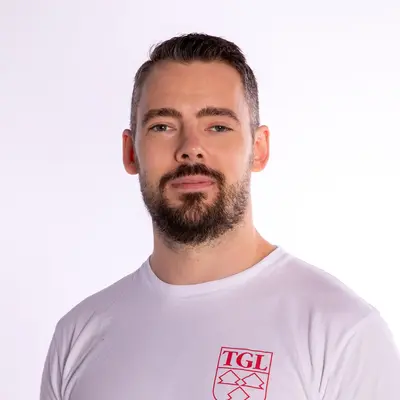Ein lächelnder Trainer im TGL-Shirt steht vor einem neutralen Hintergrund, repräsentiert den Sportverein.