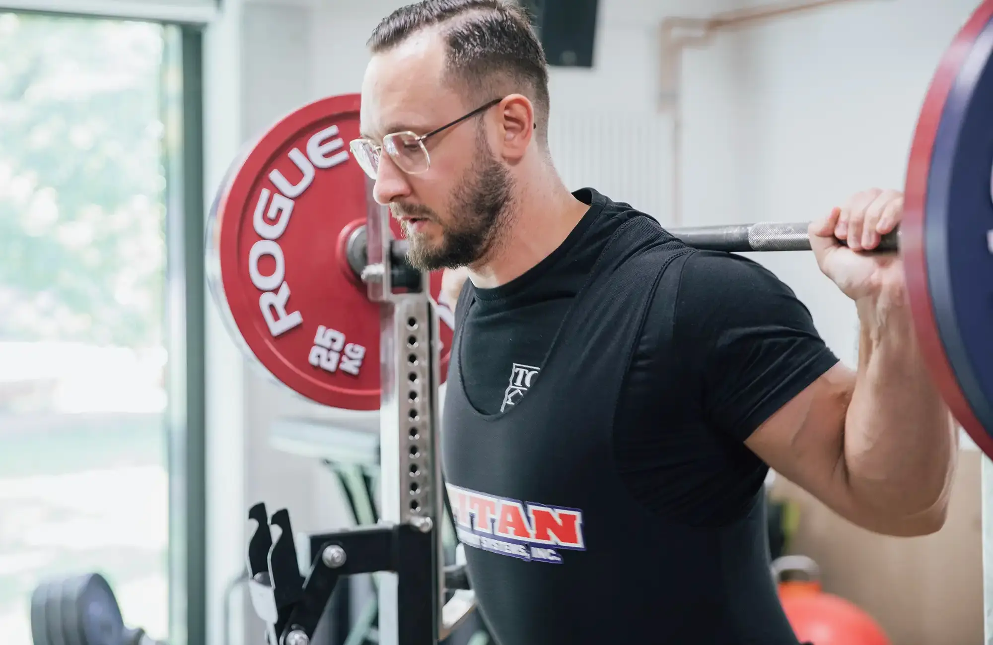 Ein Mann hebt in einem Fitnessstudio Gewichte, während er ein TGL-Shirt trägt, das seinen Sportbezug zum Verein Landshut zeigt.