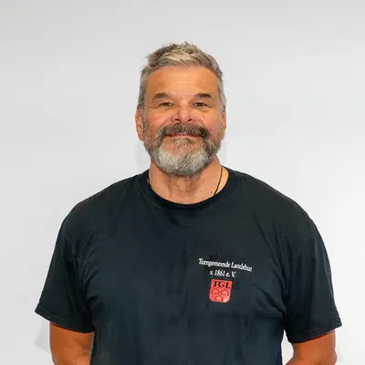 Ein lächelnder Trainer im TGL-Shirt steht vor einem neutralen Hintergrund, repräsentiert den Sportverein.