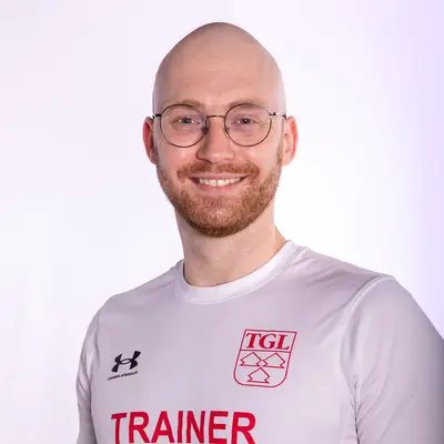 Ein lächelnder Trainer im TGL-Shirt steht vor einem neutralen Hintergrund, repräsentiert den Sportverein.
