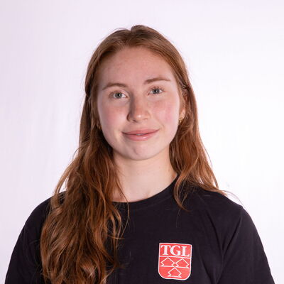 Eine junge Frau mit langen Haaren trägt ein TGL-T-Shirt und lächelt freundlich in die Kamera.