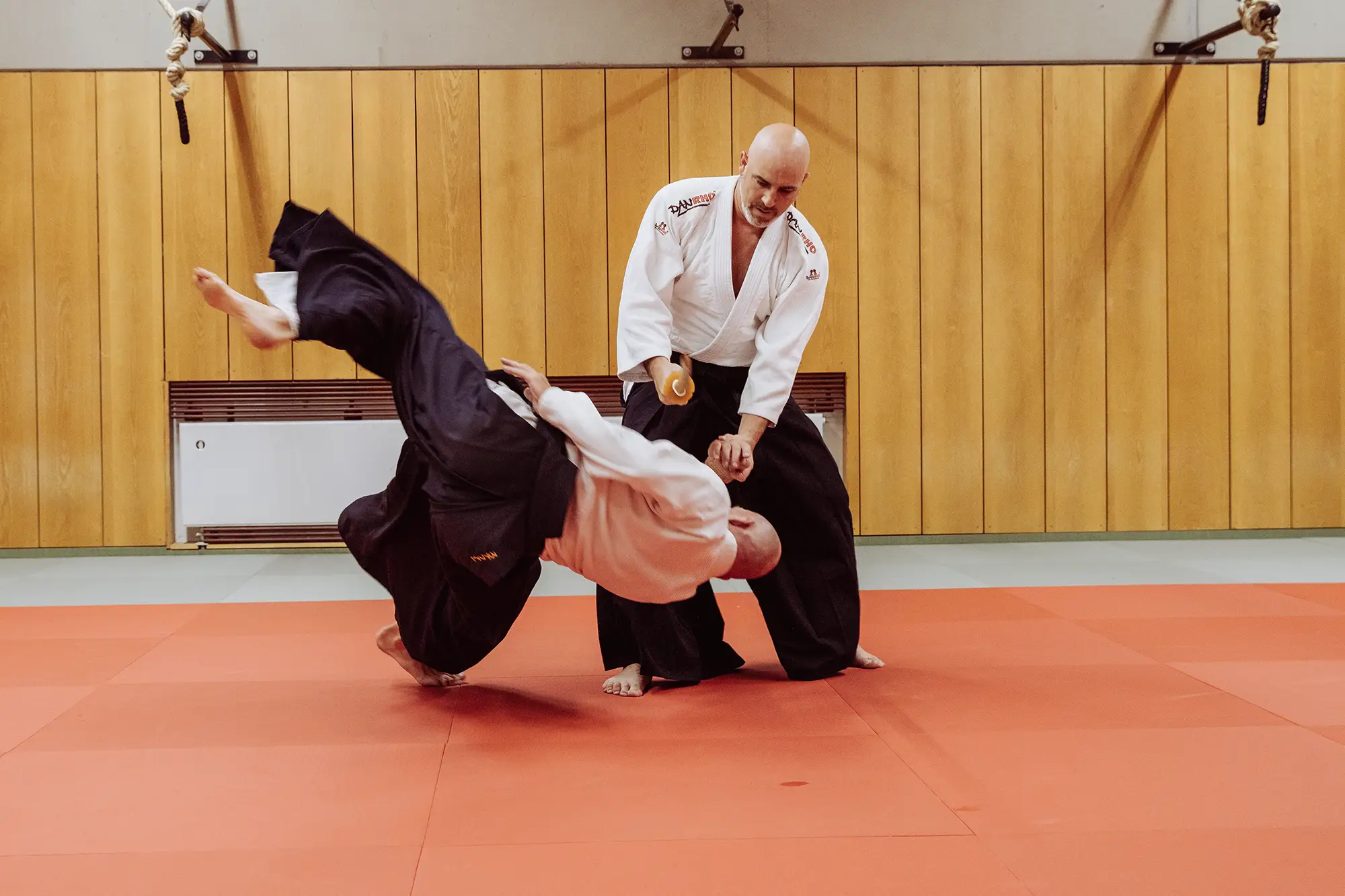 Zwei Männer üben eine Aikido-Technik in einer Turnhalle des Sportvereins Landshut auf einem roten Mattenboden.