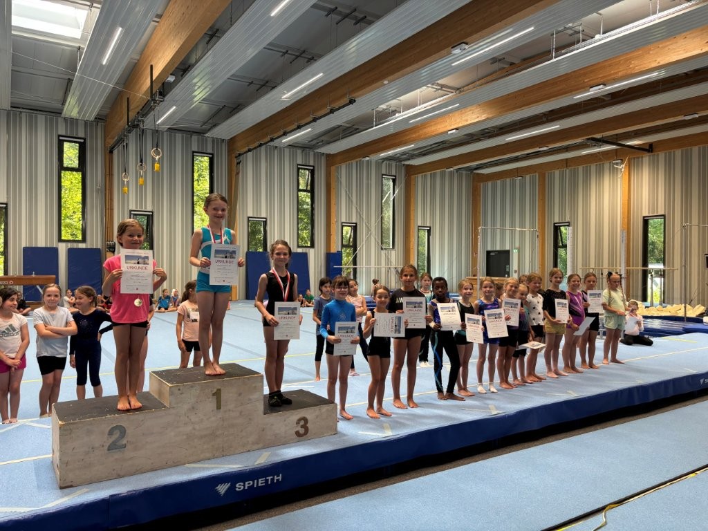 Kinder der Turngemeinde Landshut posieren stolz mit Urkunden und Medaillen auf einem Podest in einer Sporthalle.