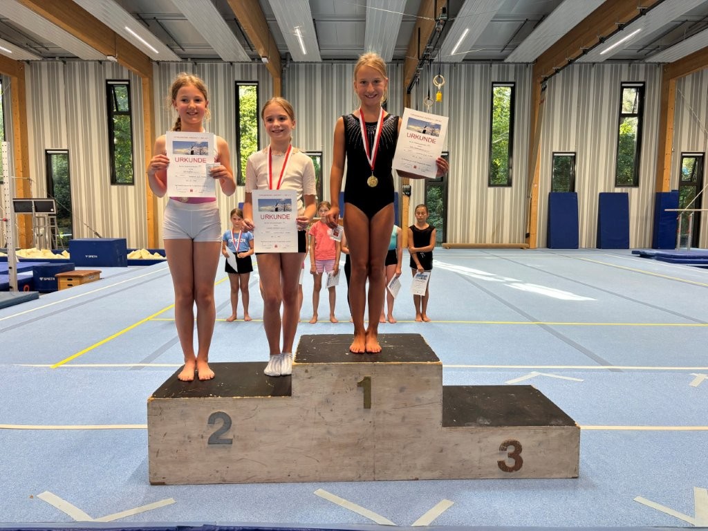 Drei junge Turnerinnen der Turngemeinde Landshut stehen stolz auf dem Siegerpodest mit Urkunden und Medaillen.