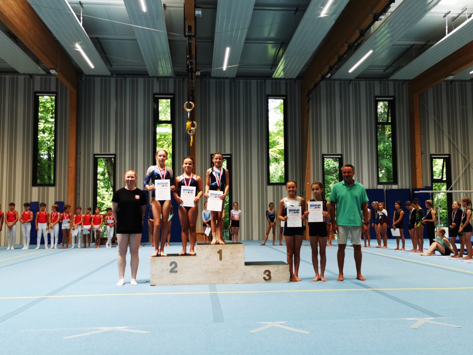 Junge Turnerinnen der Turngemeinde Landshut stehen auf dem Podest und halten stolz ihre Urkunden bei einer Vereinsmeisterschaft.