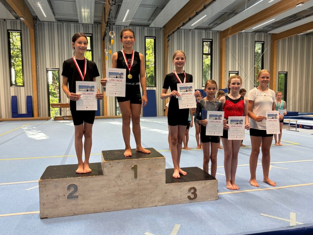 Junge Turnerinnen der Turngemeinde Landshut stehen auf dem Siegerpodest und halten stolz ihre Urkunden in der Sporthalle.