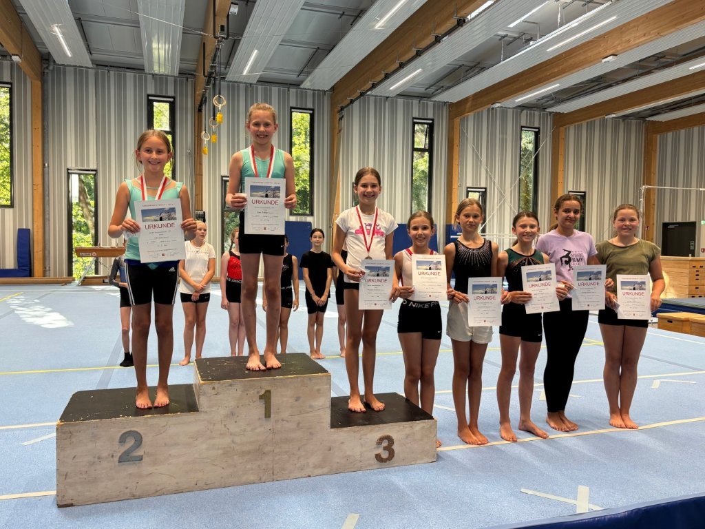 Junge Turnerinnen des Vereins Landshut präsentieren stolz ihre Urkunden auf einem Siegerpodest in einer Sporthalle.